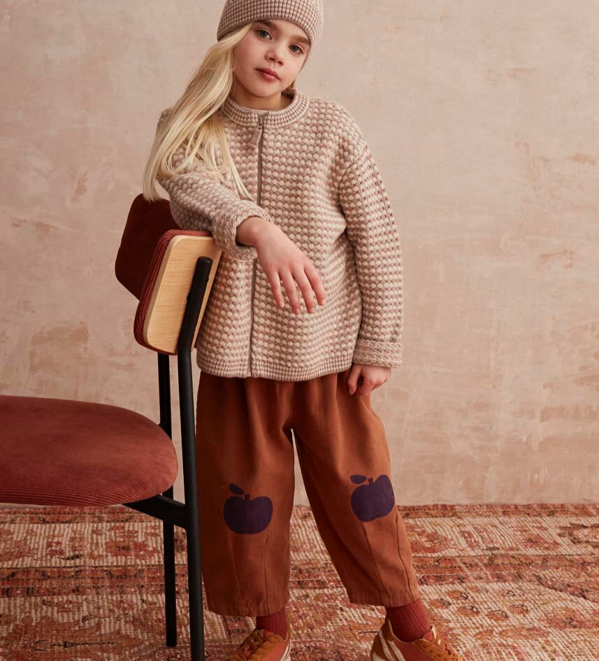 Baby in a purple sweater and brown pants standing next to an adult on a patterned rug.