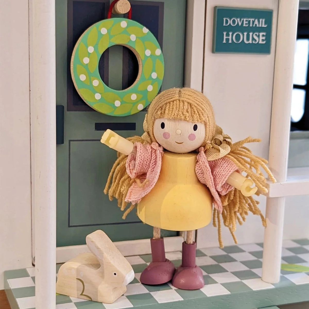 Toy doll with a rabbit figurine on a checkered platform with a green wreath and 'Dovetail House' sign in the background.