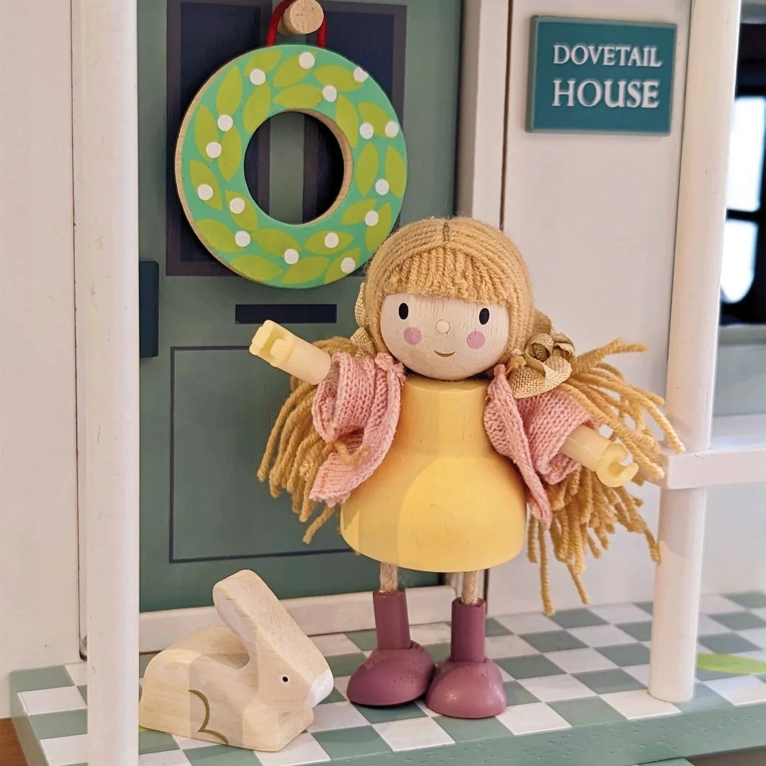 Toy doll with a rabbit figurine on a checkered platform with a green wreath and 'Dovetail House' sign in the background.