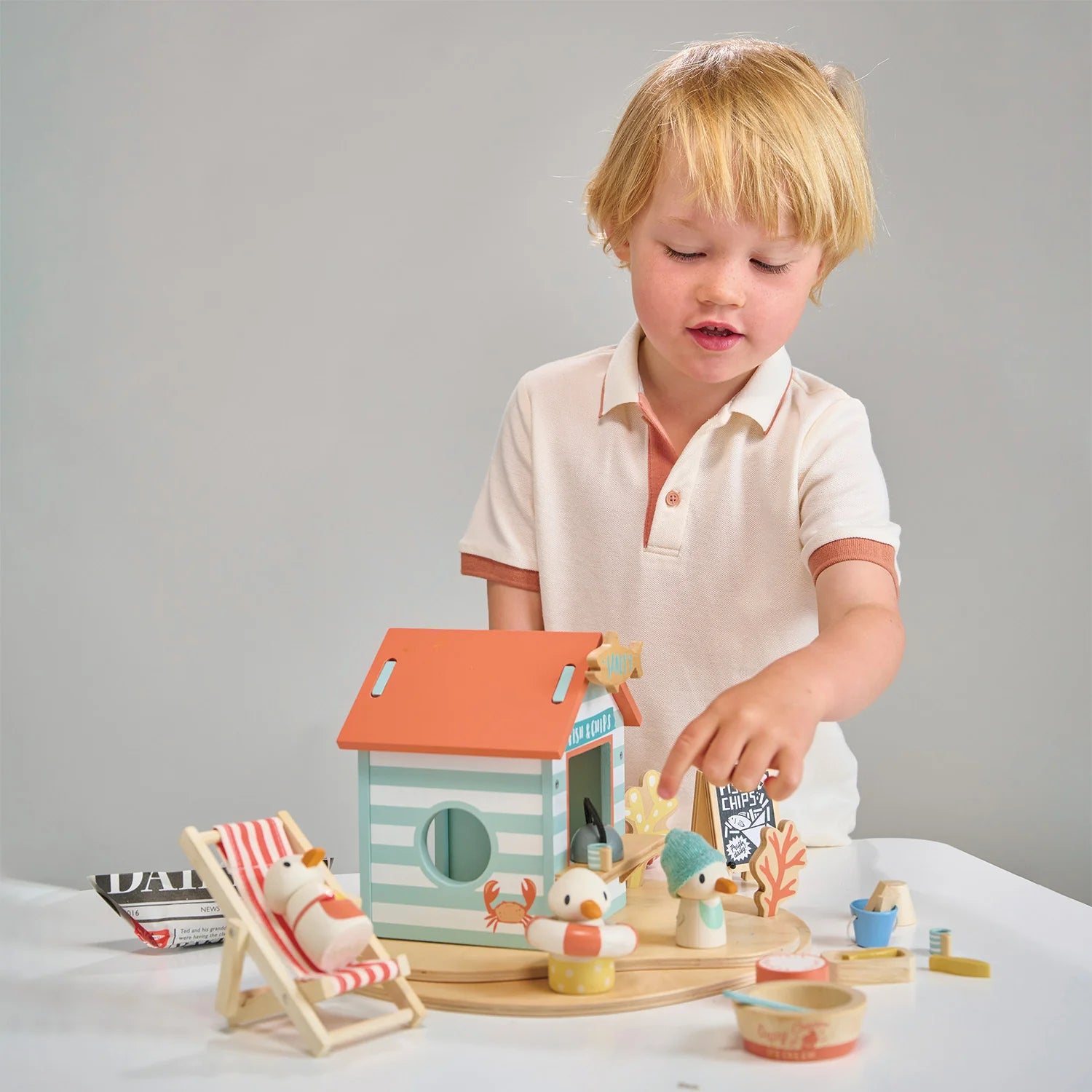 Child playing with a wooden toy beach house set on a white surface.