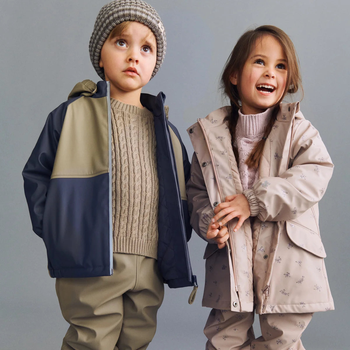 Two children wearing stylish jackets and hats against a gray background