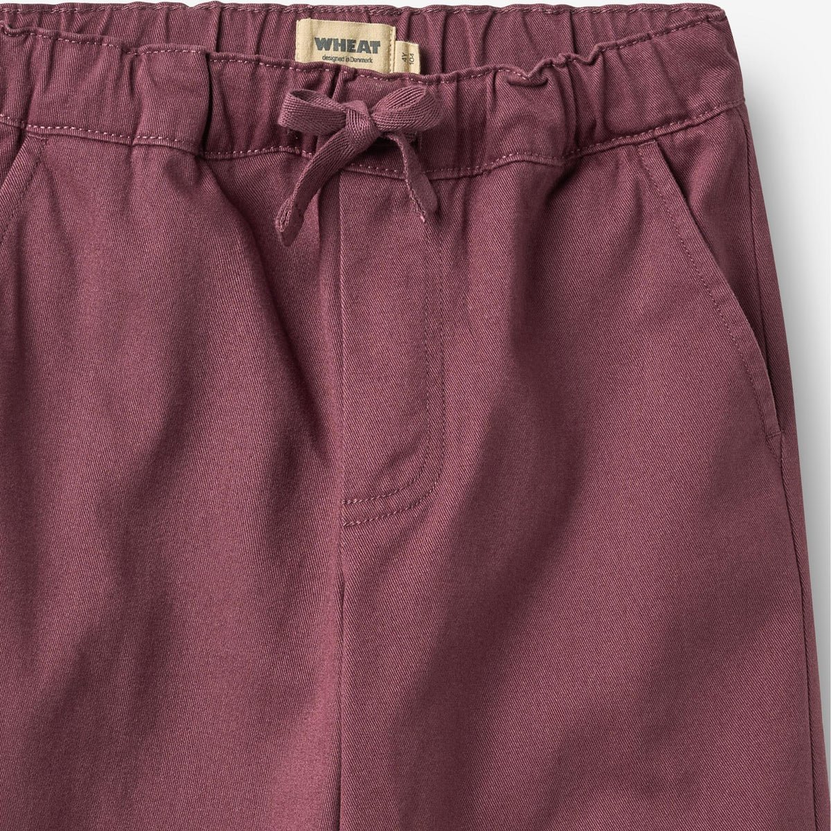 Close-up of a pair of dusty rose drawstring pants showing the elastic waistband, a beige woven label reading 'WHEAT', and the side pocket seam.