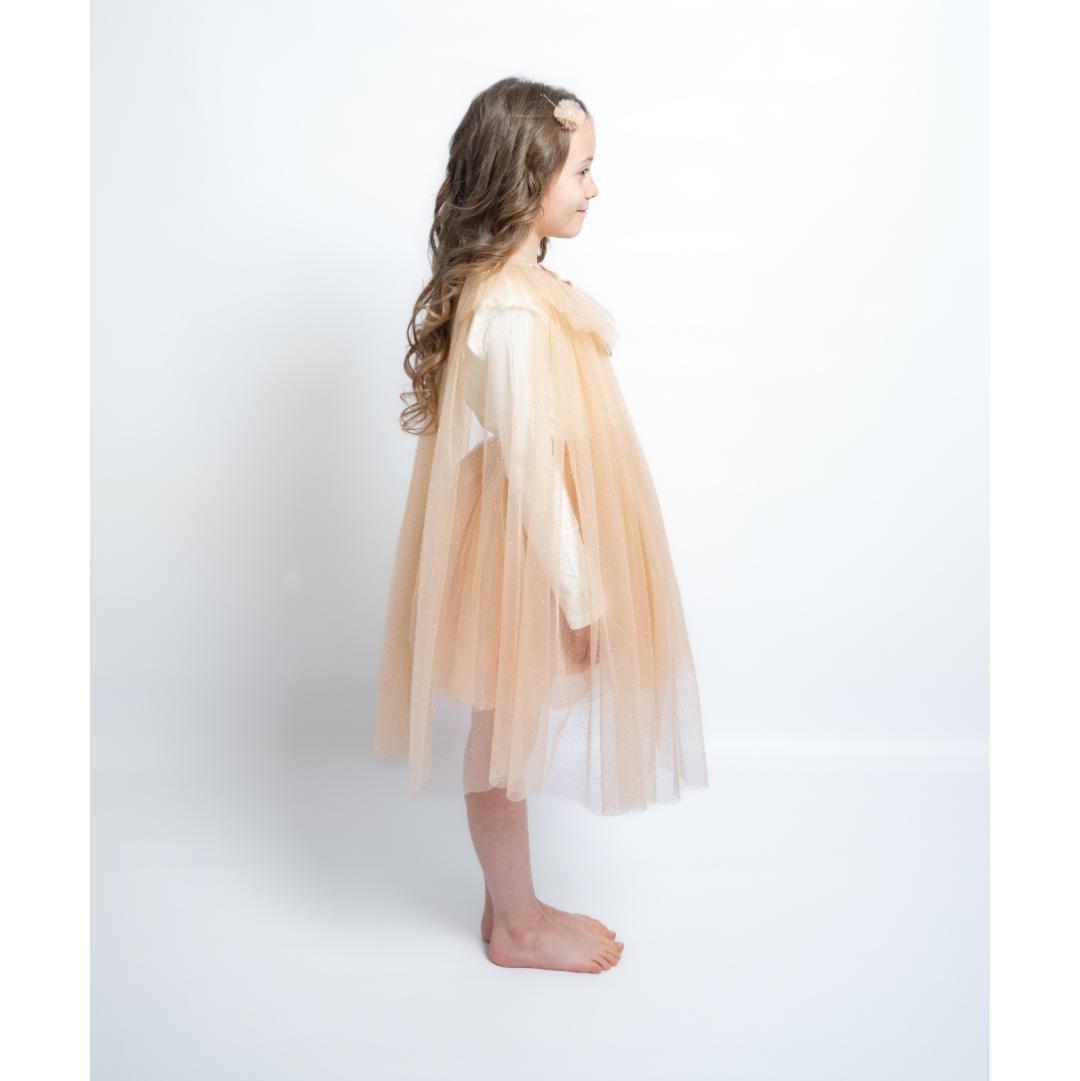 A young girl with long wavy brown hair stands in profile wearing a light peach tulle cape over a white long-sleeve top, barefoot against a plain white studio background.