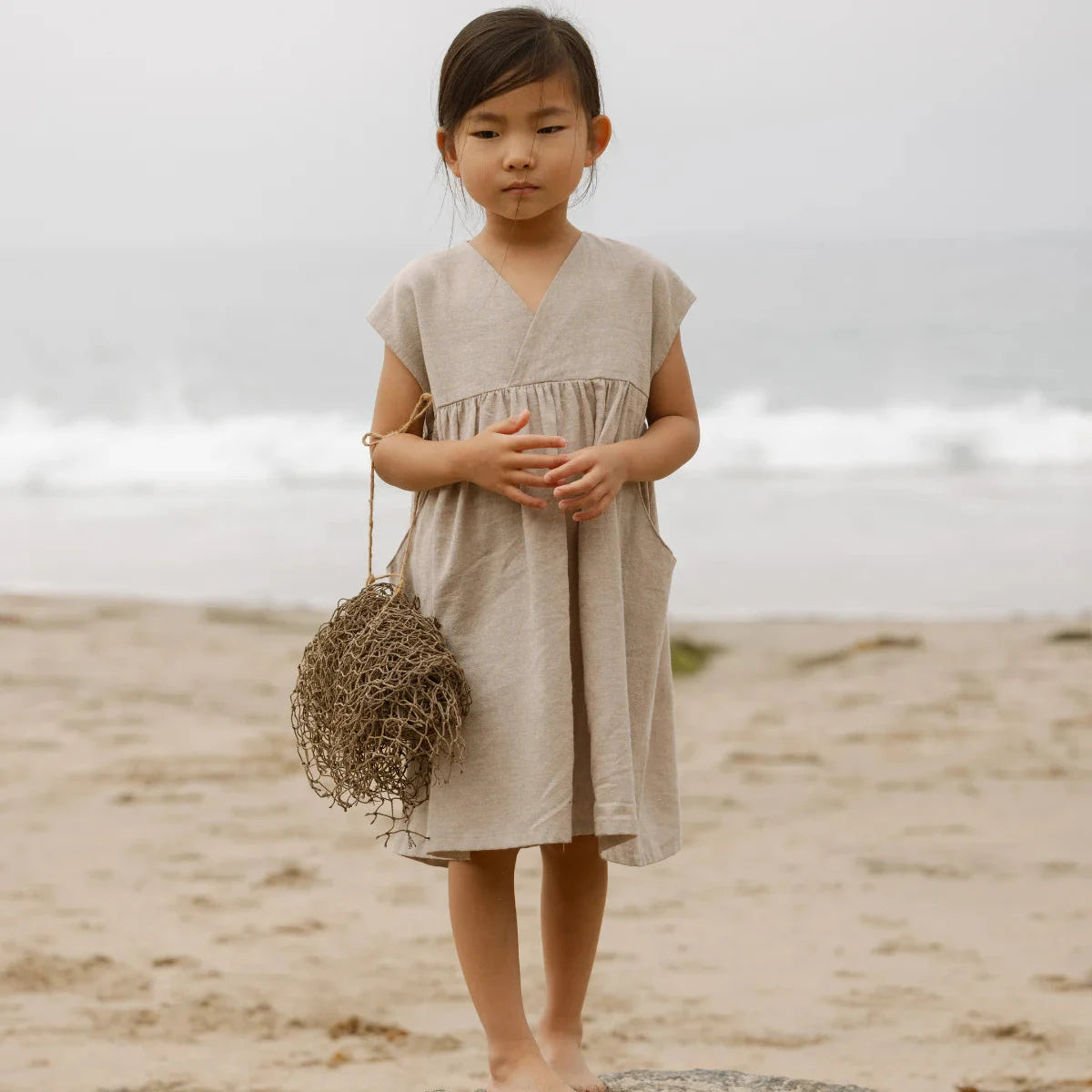 Young girl in a beige dress standing on a beach holding a bundle of dried grass.