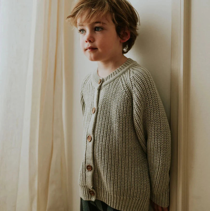 Child wearing a knitted cardigan standing against a white wall with curtains.