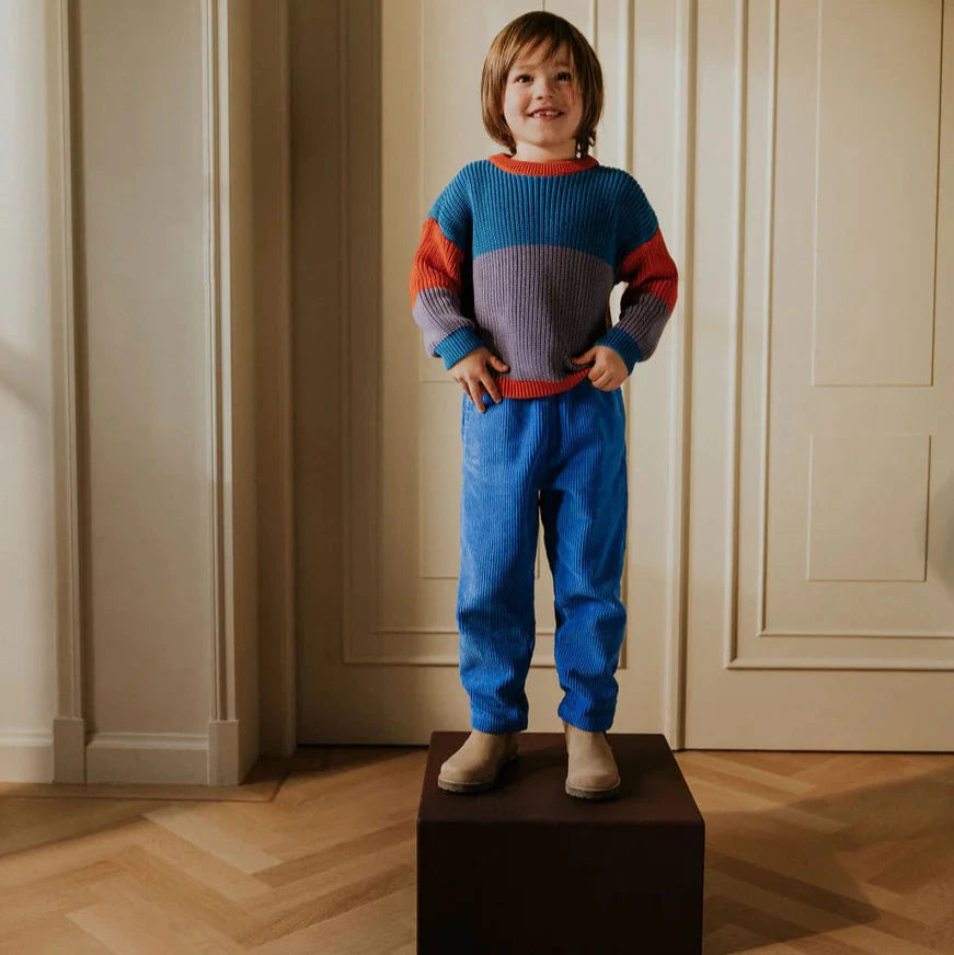 Child standing on a box wearing a color block sweater and blue cord pants with wooden flooring and white walls