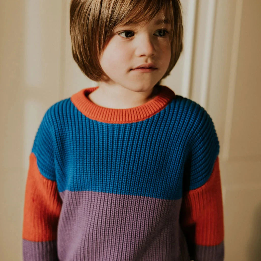Child standing on a box wearing a color block sweater and blue cord pants with wooden flooring and white walls
