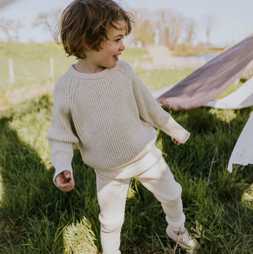Child in a cream sweater and cream pants standing in a grassy field with a tent in the background.