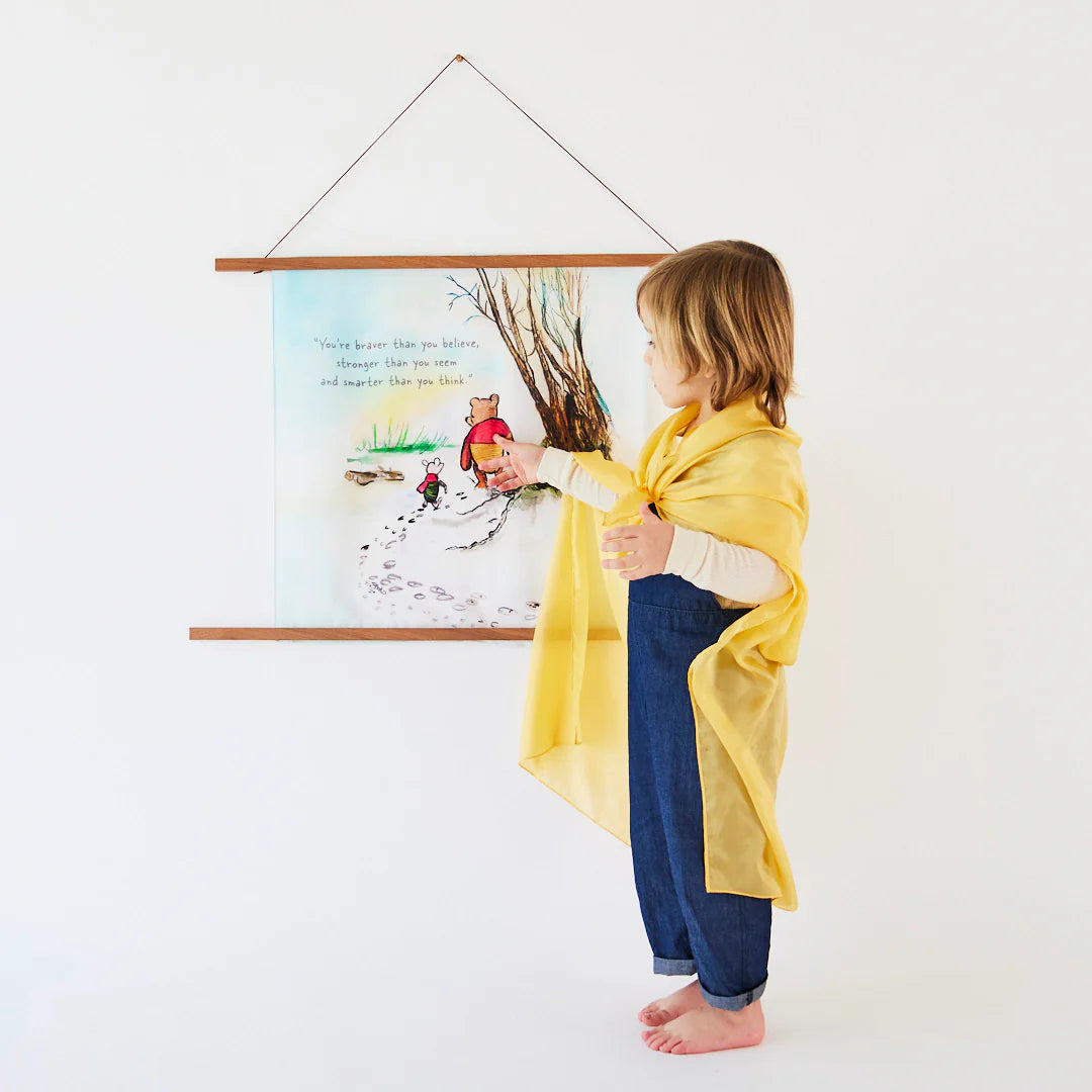 Child in a yellow coat standing next to a wall with a framed picture of a snowy scene with characters.