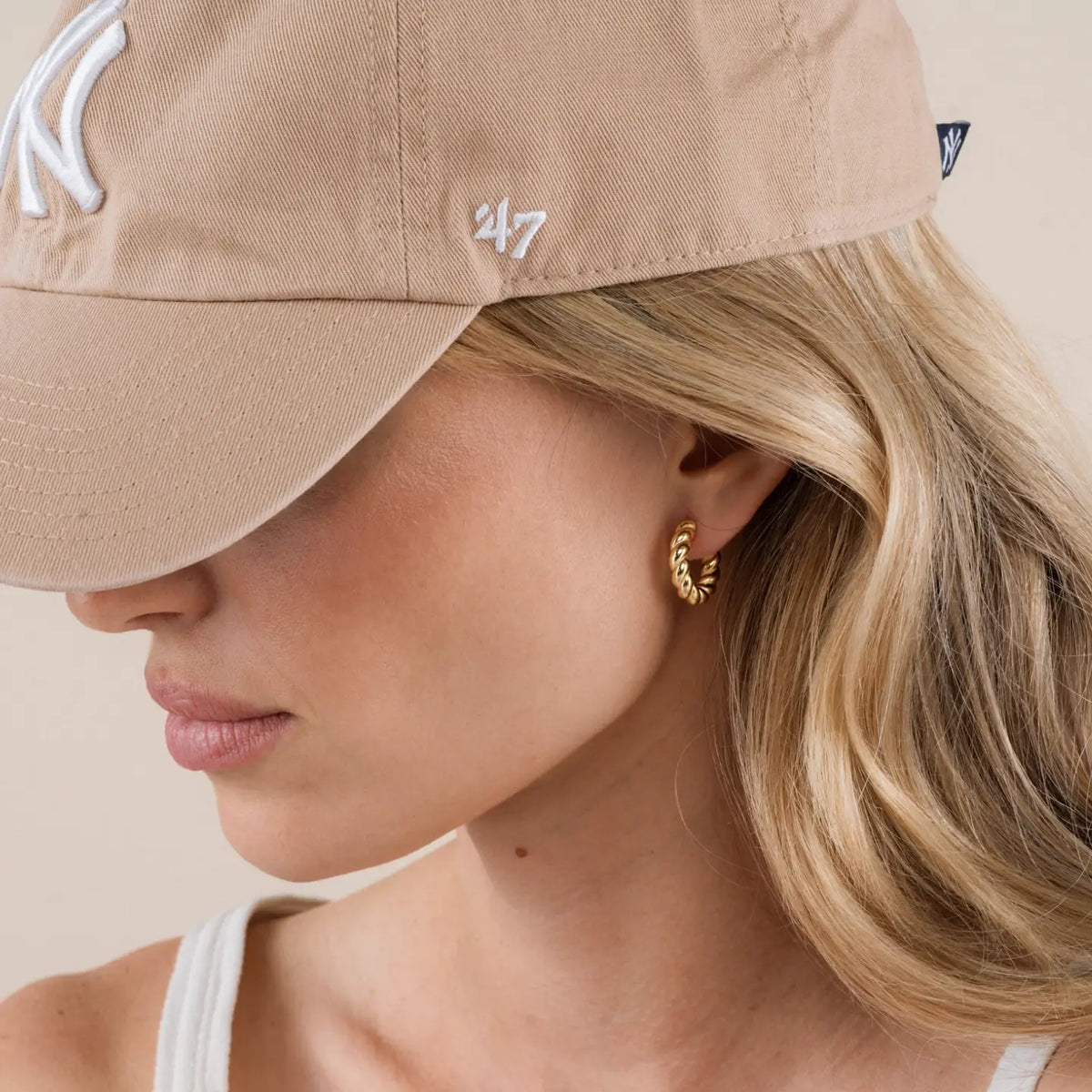 Close-up of a woman wearing a beige cap with a logo, gold hoop earrings, and a white top.