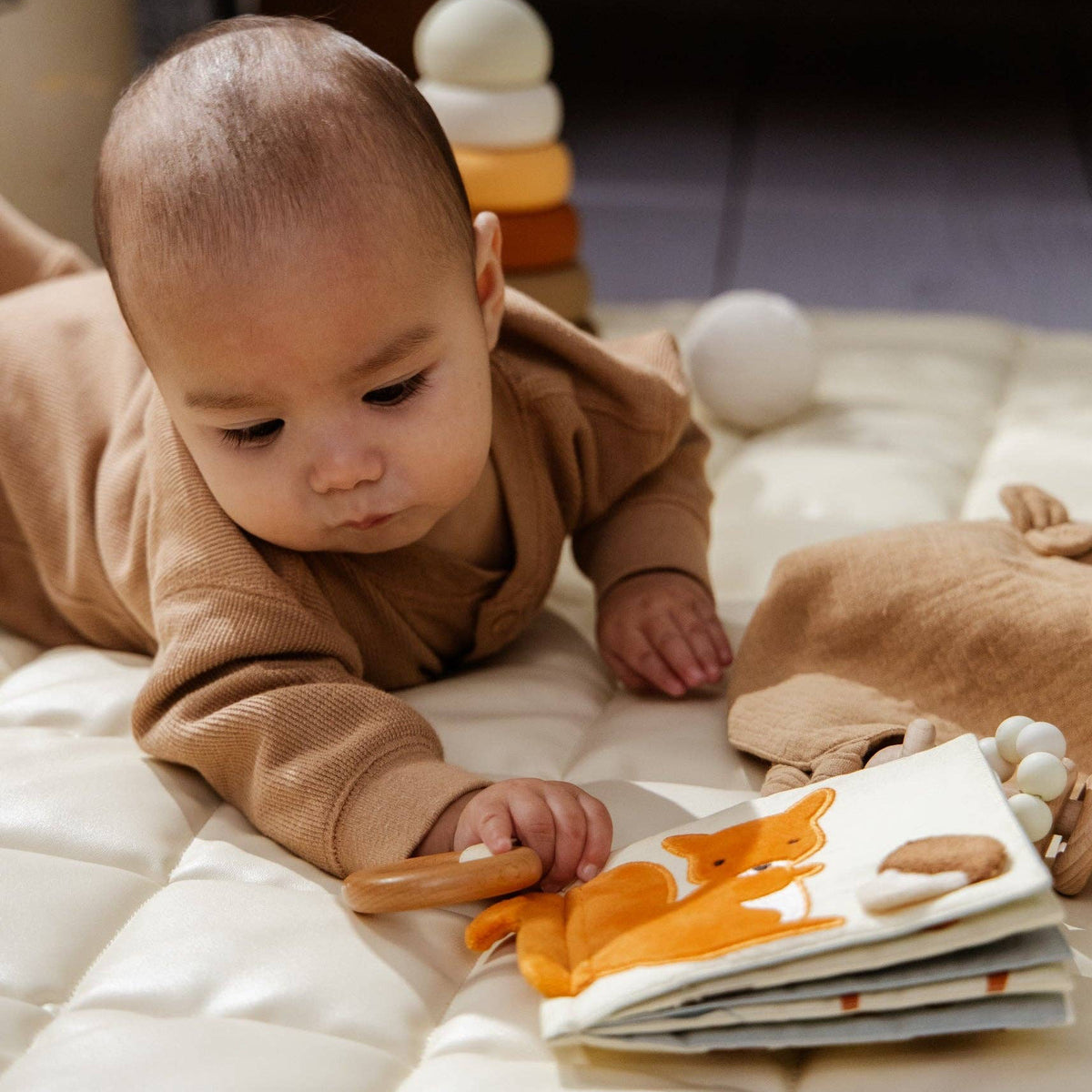 Baby lying on a white blanket with toys around, holding a wooden rattle.