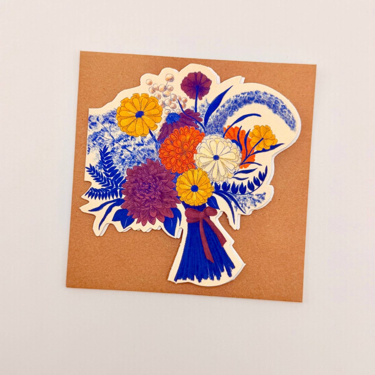 A square kraft paper card featuring a colorful floral bouquet illustration in blue, yellow, orange, purple, and white, photographed against a plain off-white background.