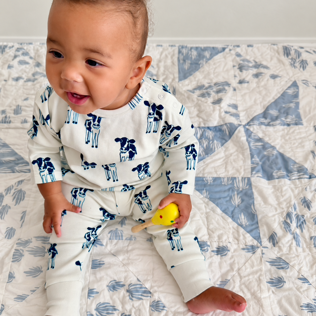 A baby wearing a white long-sleeve pajama set with a blue cow print sits on a blue and white quilted blanket, holding a small yellow toy.