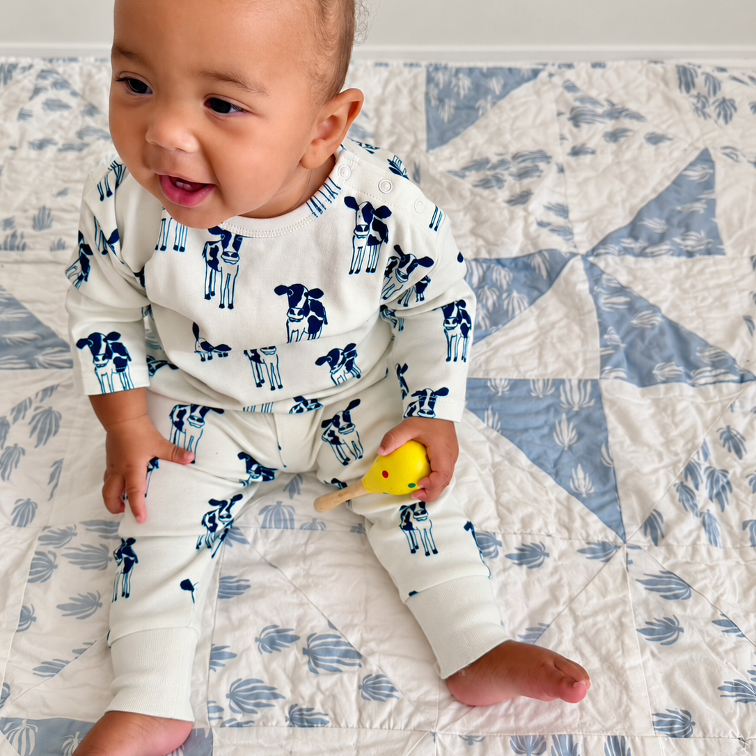 A baby wearing a white long-sleeve pajama set with a blue cow print sits on a blue and white quilted blanket, holding a small yellow toy.