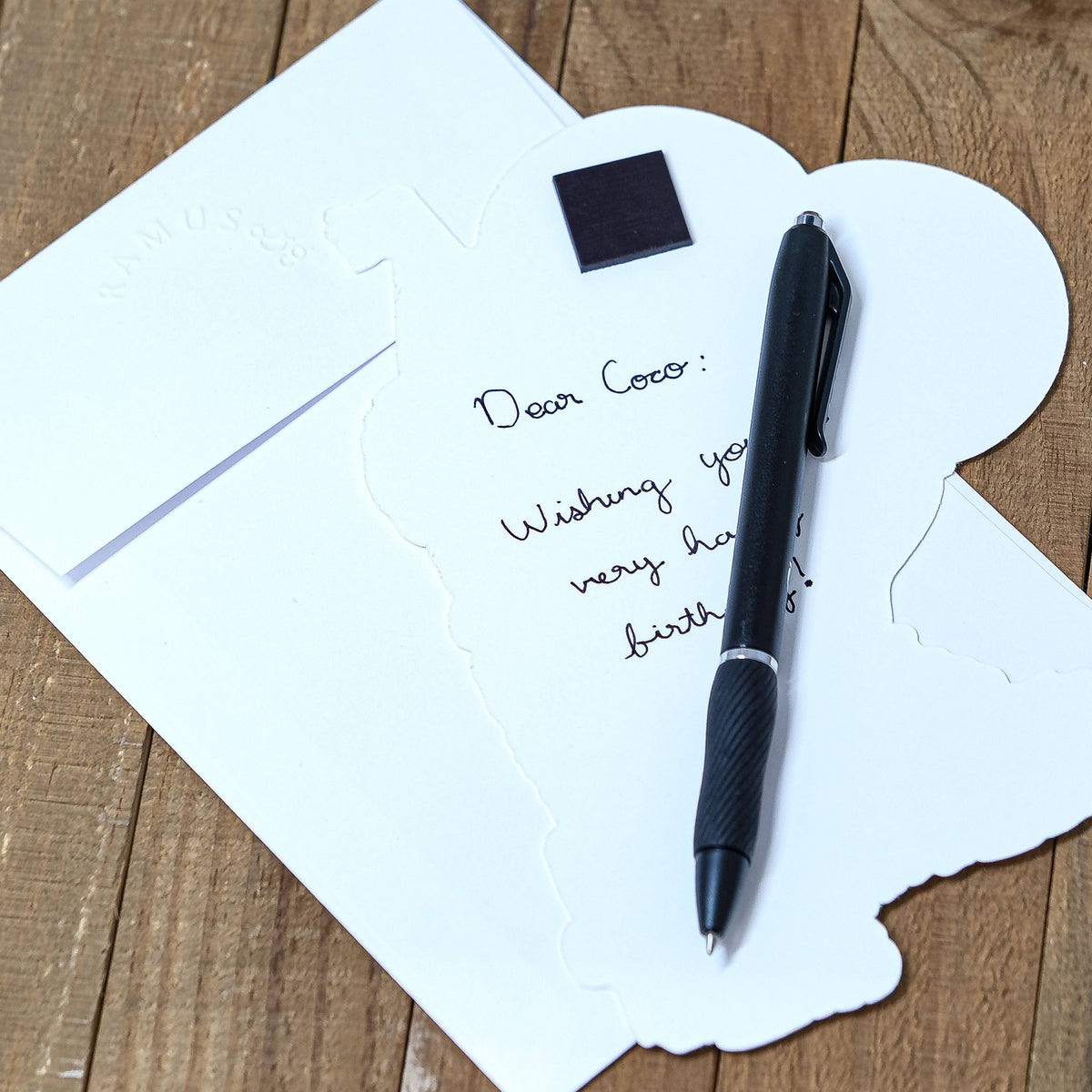 A white birthday card with a handwritten message, shown on a wooden surface alongside a black pen and a white envelope.