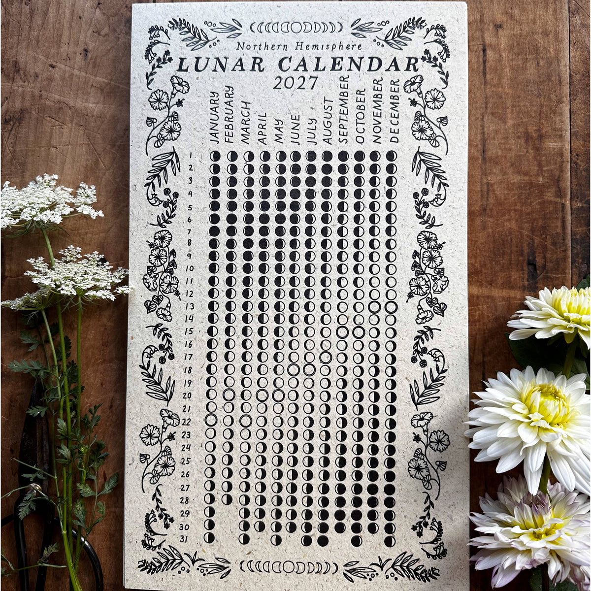 A rectangular paper lunar calendar for 2027 laid flat on a rustic wooden surface, surrounded by white flowers. The calendar has a cream background with black printed text, moon phases, and decorative border illustrations.