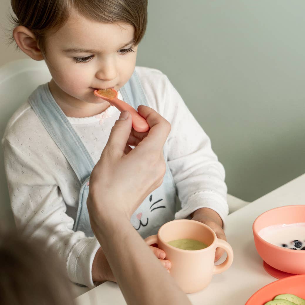 Child being fed by an adult with a spoon, sitting at a table with a cup and bowl.