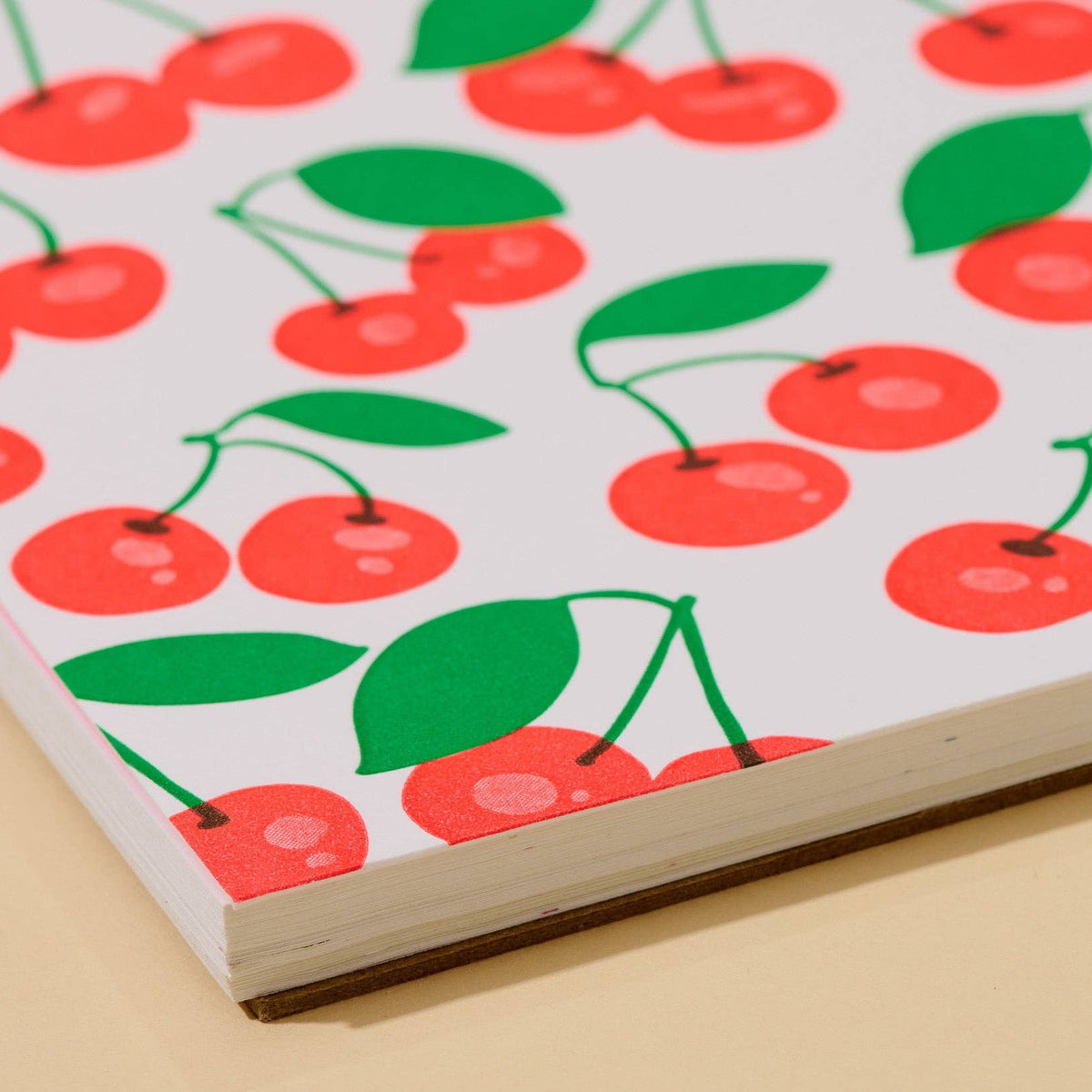 A close-up of a notebook or book cover featuring a repeating cherry print on a white background, photographed at an angle on a light beige surface.