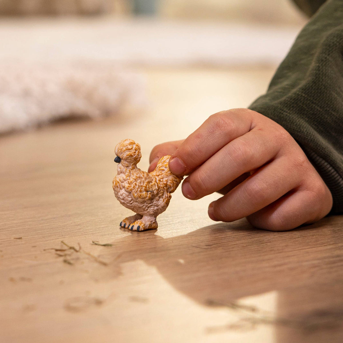 A small decorative chicken figurine standing on a wooden floor, with a child's hand reaching toward it and a dark green sleeve visible.
