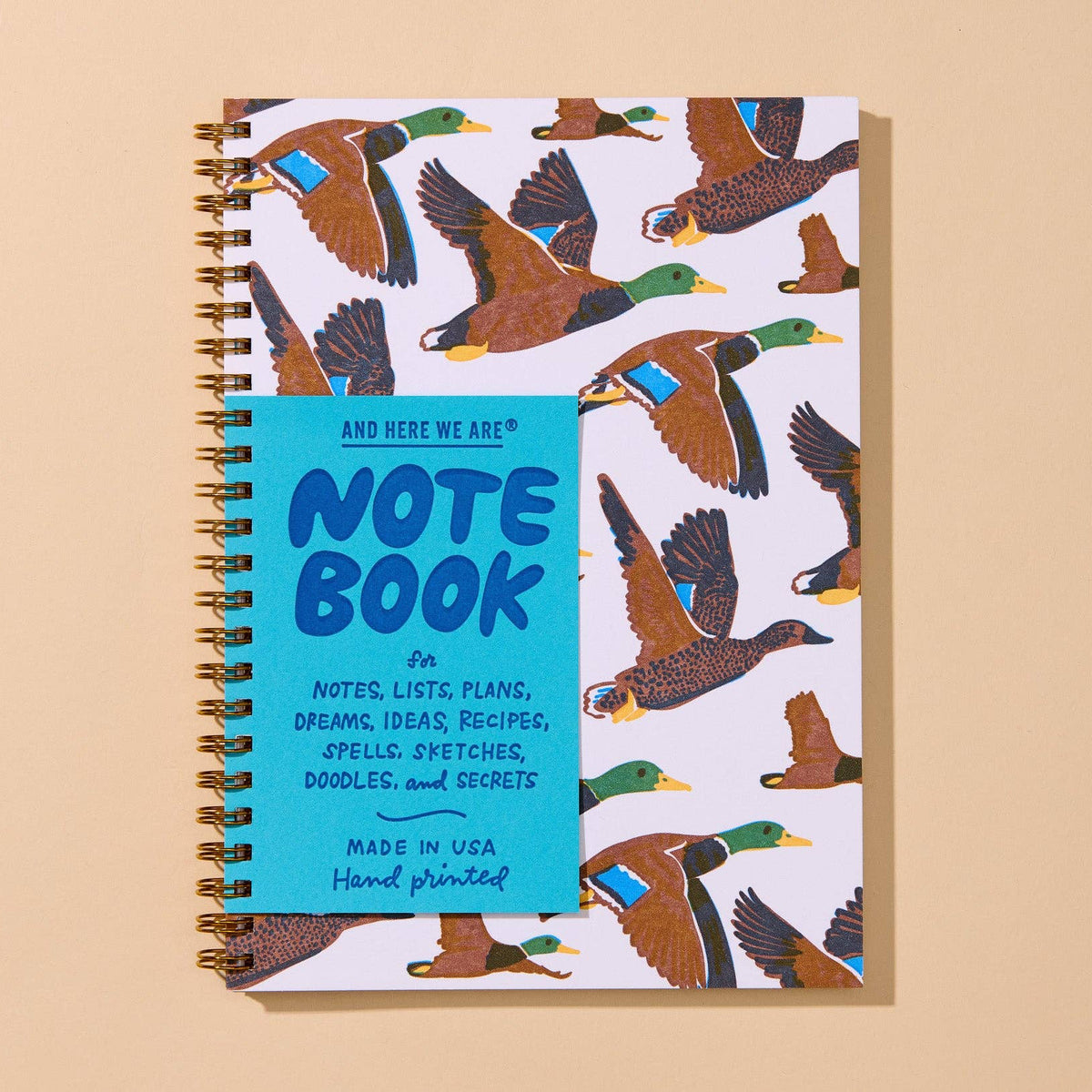 A spiral-bound notebook with a white cover printed with a pattern of flying mallards in brown, green, yellow, and blue, featuring a bright turquoise blue title block on the front that reads 'NOTE BOOK'. The notebook is photographed from above on a solid beige background.