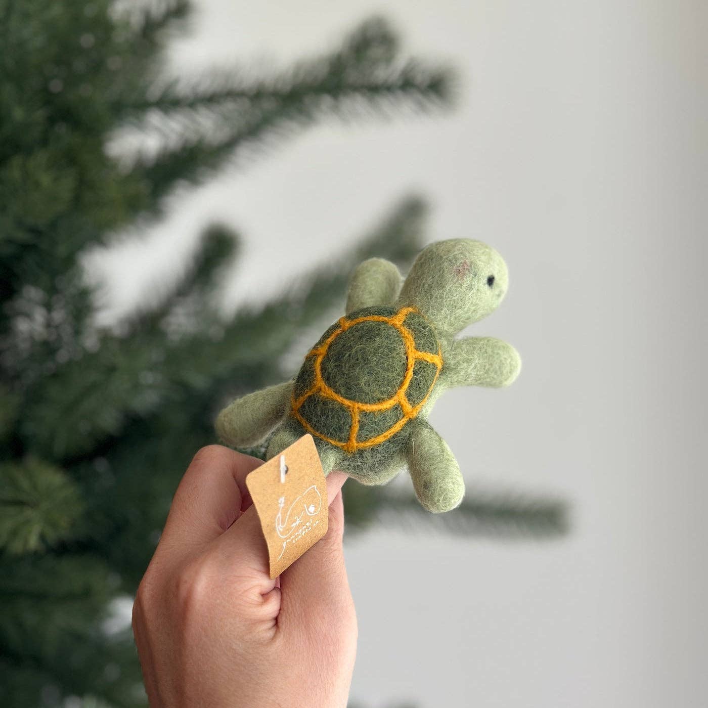 A hand holds a small felted wool turtle ornament against a soft-focus background of a Christmas tree and a light gray wall. The turtle has a light green body and a darker green shell with orange felted lines.
