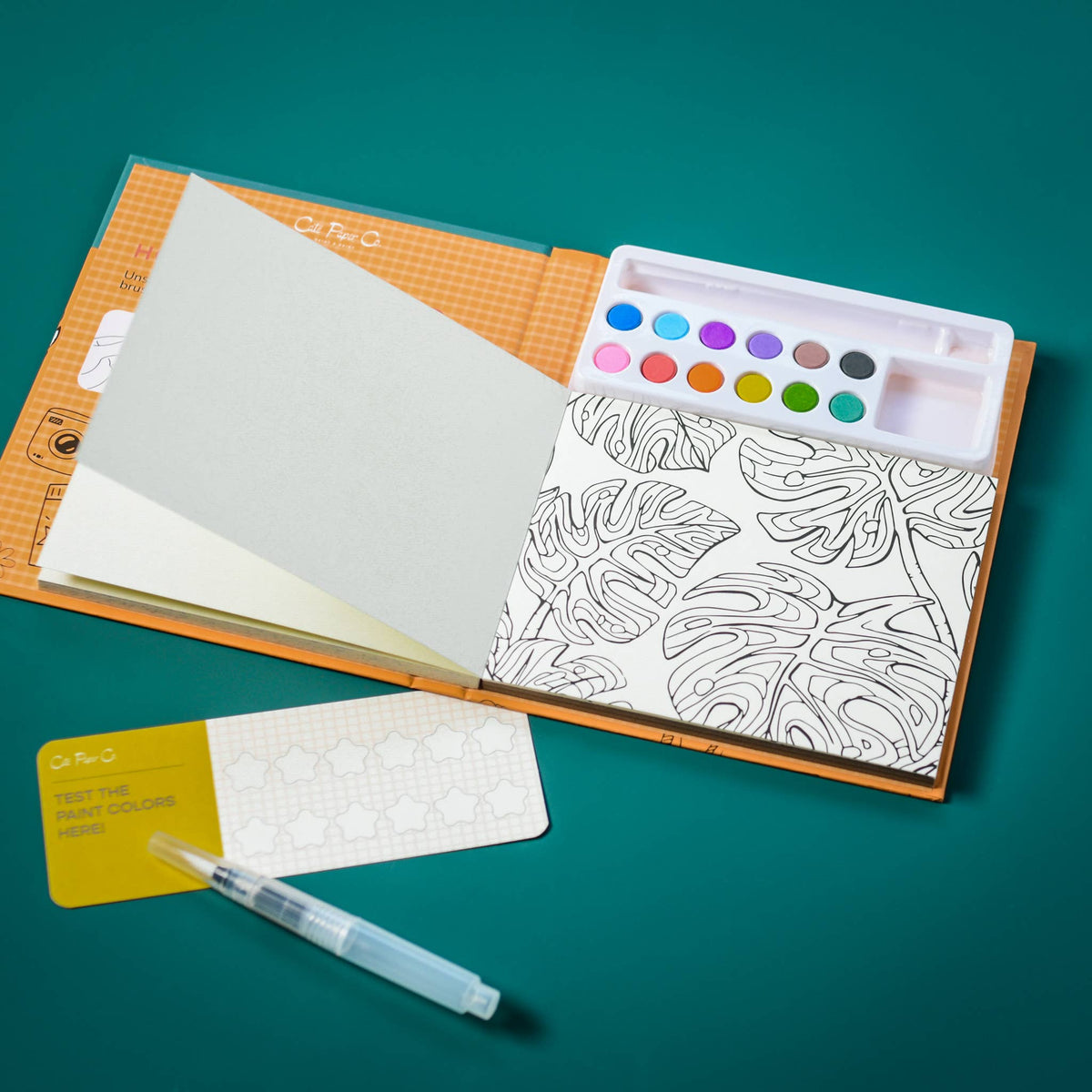 Open notebook with watercolor paint set and pen on a teal background
