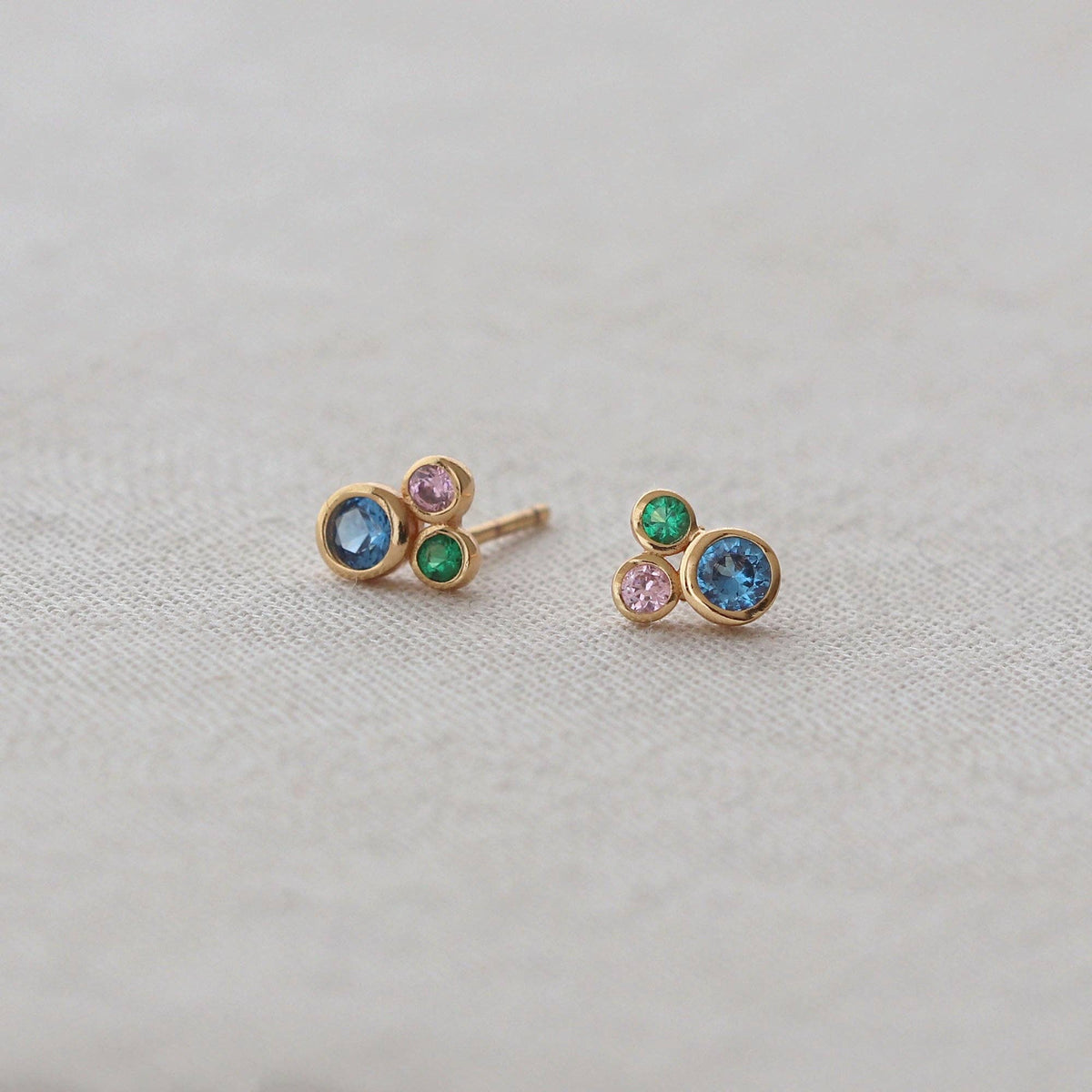 A pair of gold stud earrings with small round gemstones in blue, green, and pink, displayed on a textured beige fabric background.