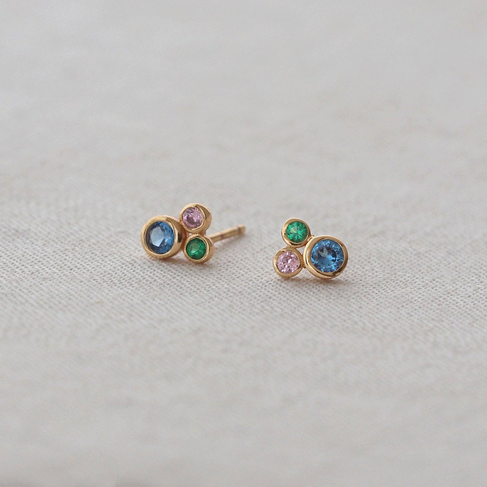 A pair of gold stud earrings with small round gemstones in blue, green, and pink, displayed on a textured beige fabric background.
