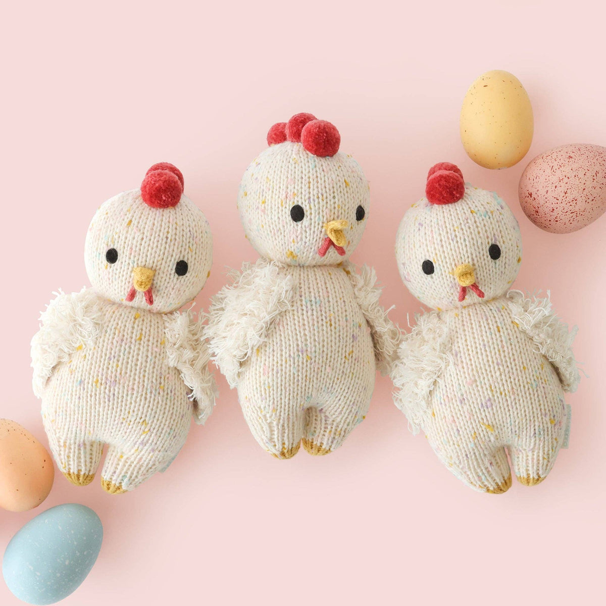 Three knitted chicken toys with colorful eggs on a pink background