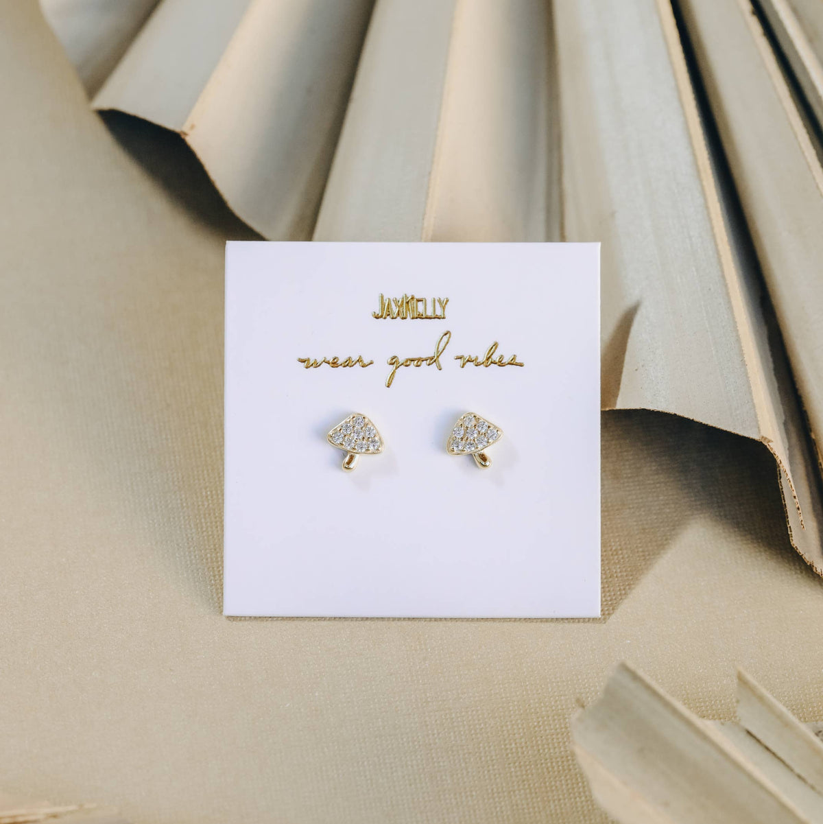 Gold mushroom-shaped earrings on a white card with 'JaxKelly branding, against a beige textured background.