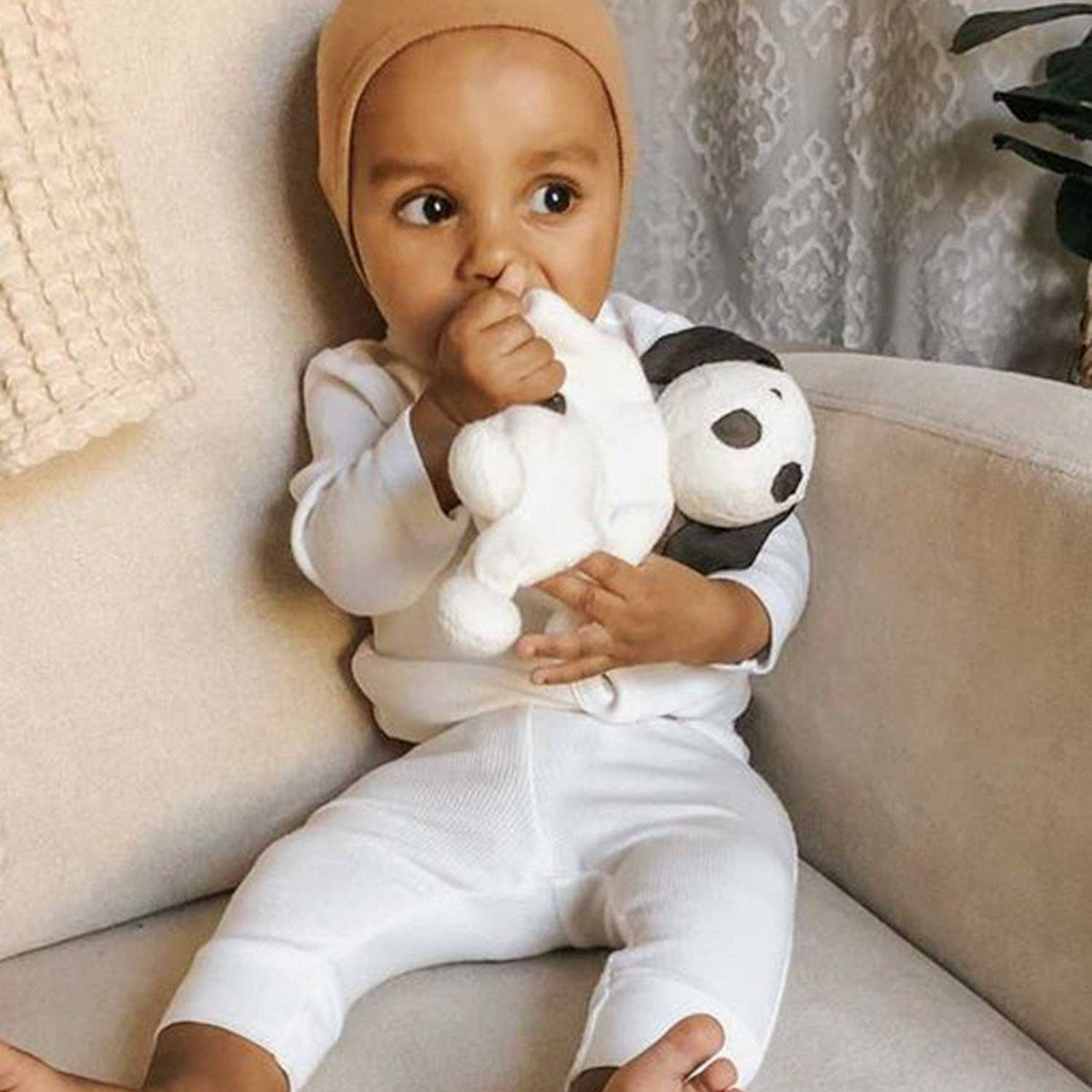 Baby in a white outfit with a beige hat  holding a toy on a couch.