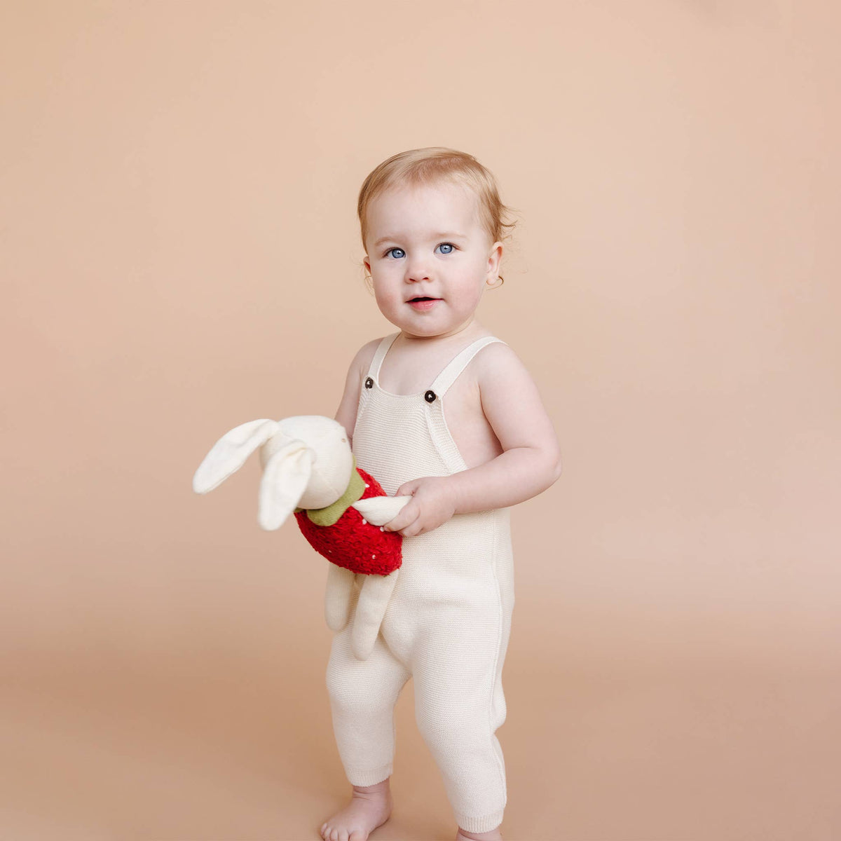 Baby in white overalls holding a plush toy against a beige background