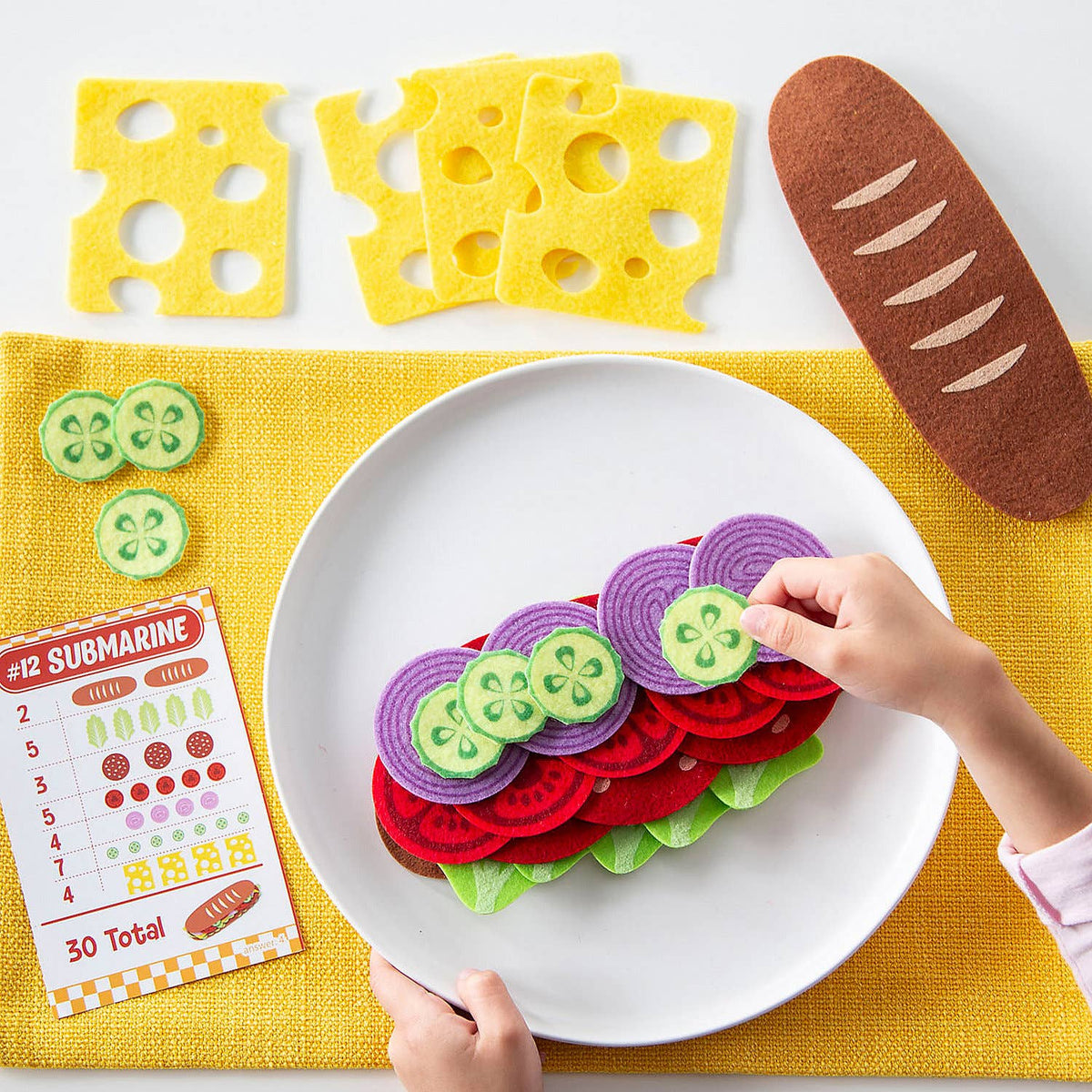 Colorful felt submarine sandwich set on a white plate with felt cheese, bread, and vegetables on a yellow mat.