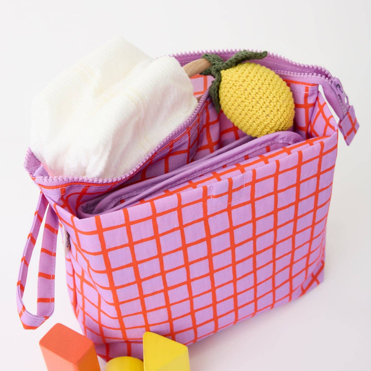 A pink and orange checkered fabric diaper bag with a purple zipper, shown open with a white cloth and a yellow crocheted pineapple toy inside. Three small colorful blocks are placed in front on a white background.