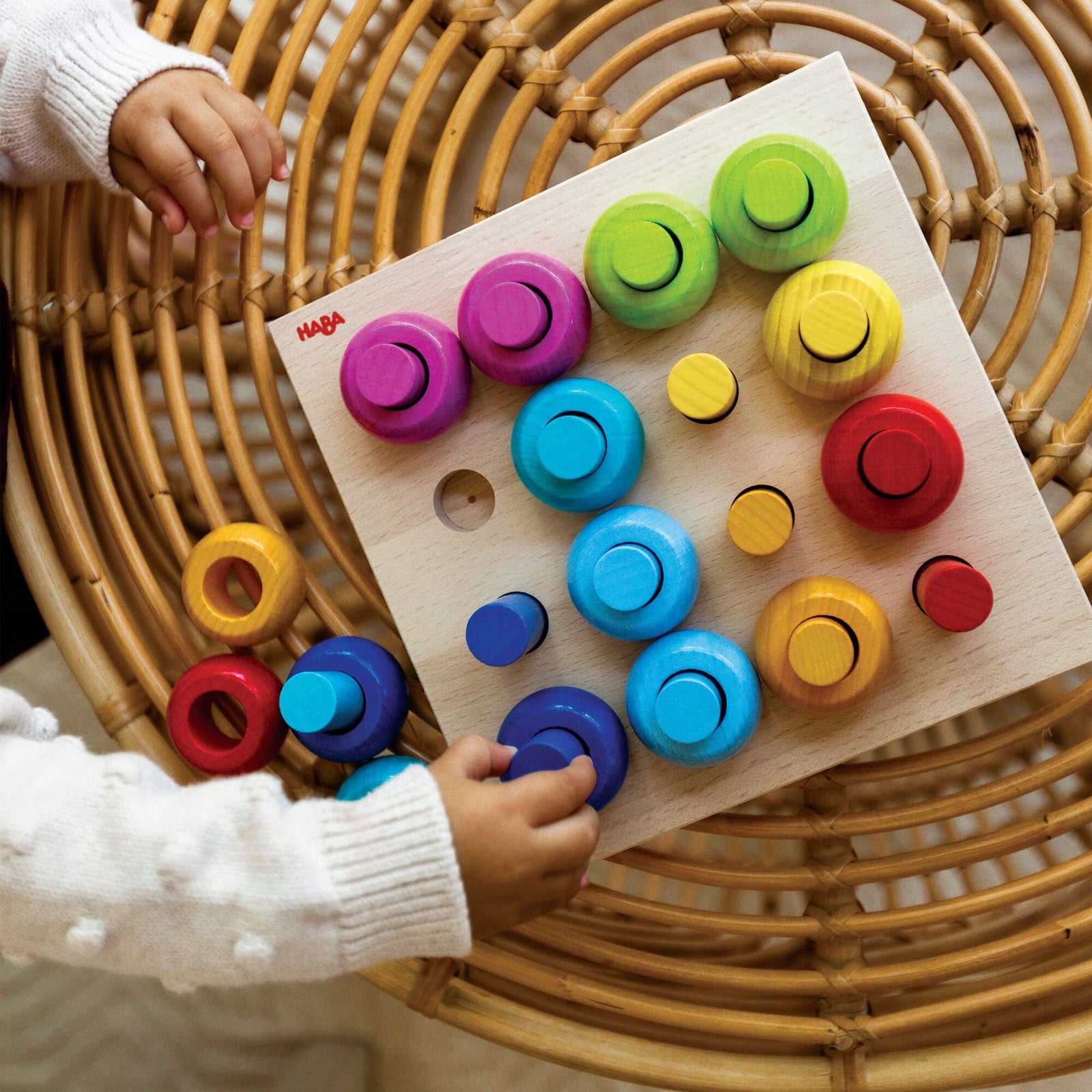 Colorful wooden puzzle toy on a wicker surface with child's hands interacting with it.