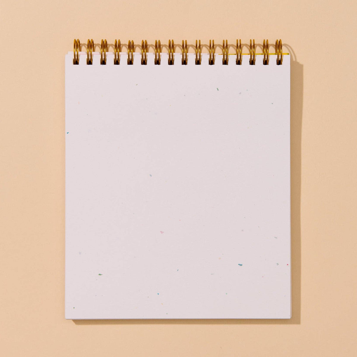 A blank white spiral-bound notepad with a gold wire binding, standing upright against a solid warm beige background.