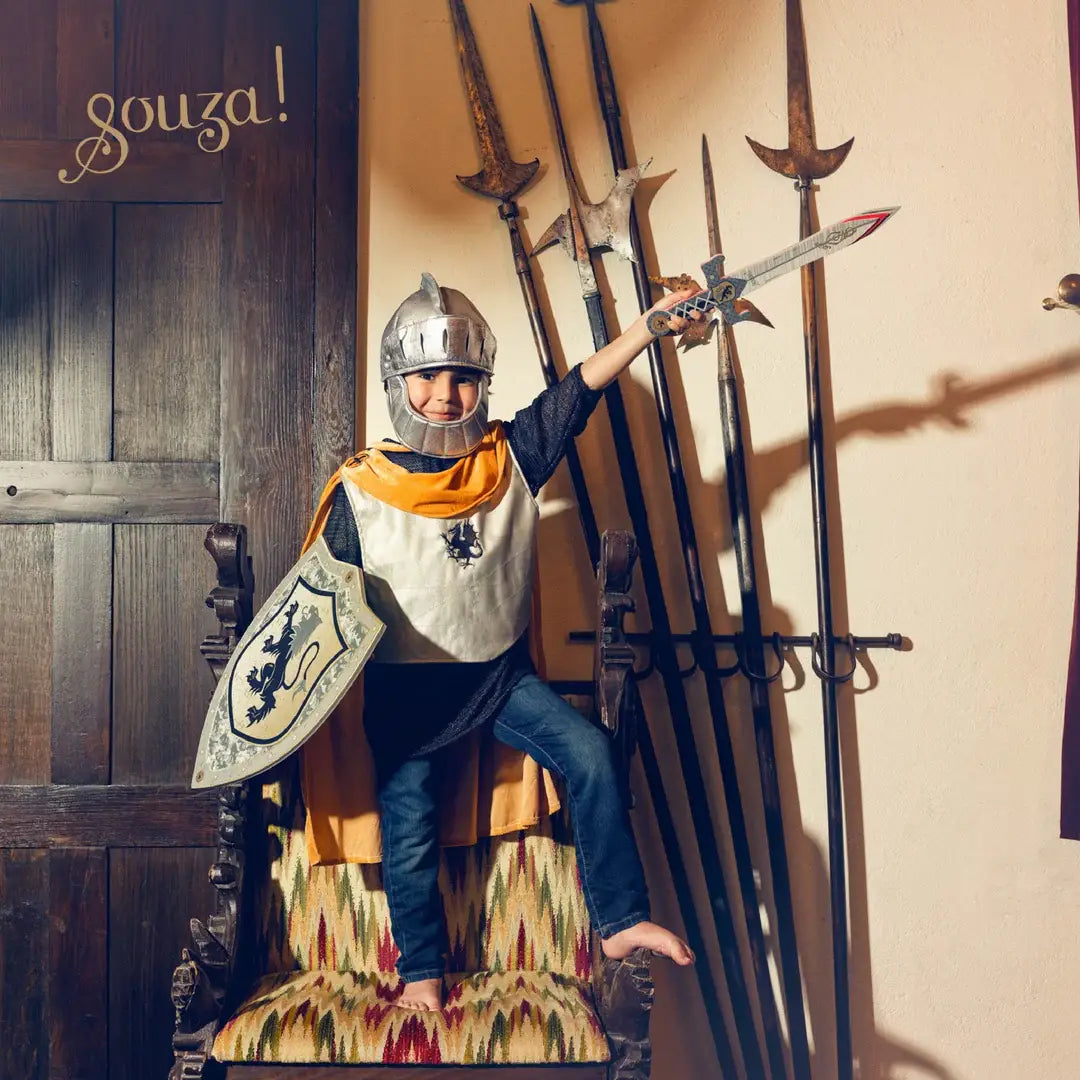 Child in knight costume holding a sword in front of various weapons on a wall.