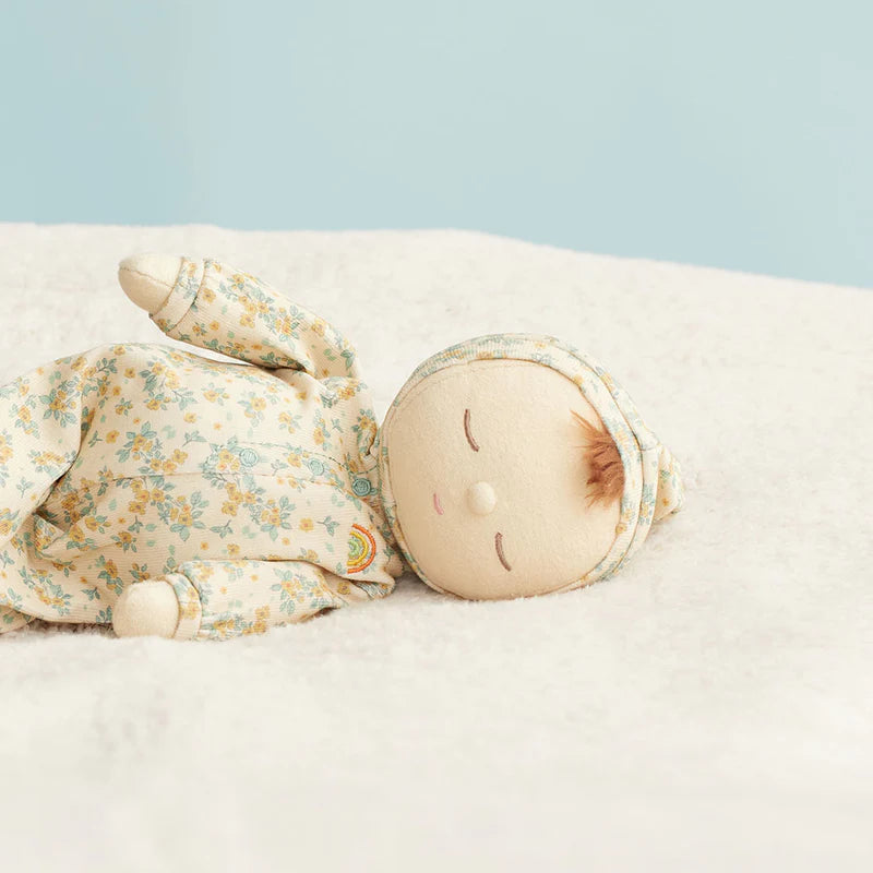 A sleeping fabric doll with a floral-patterned bodysuit and bonnet lies on a soft white blanket against a light blue background.