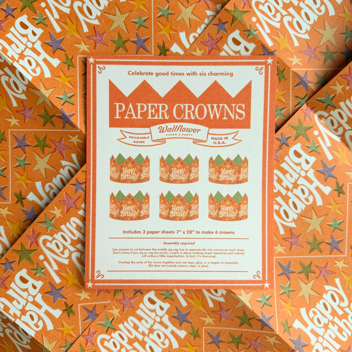 A product package for 'Paper Crowns' by Wallflower, laid flat on top of two matching orange patterned paper sheets. The package is cream and orange with illustrations of six paper crowns, and the background sheets feature a colorful star pattern.