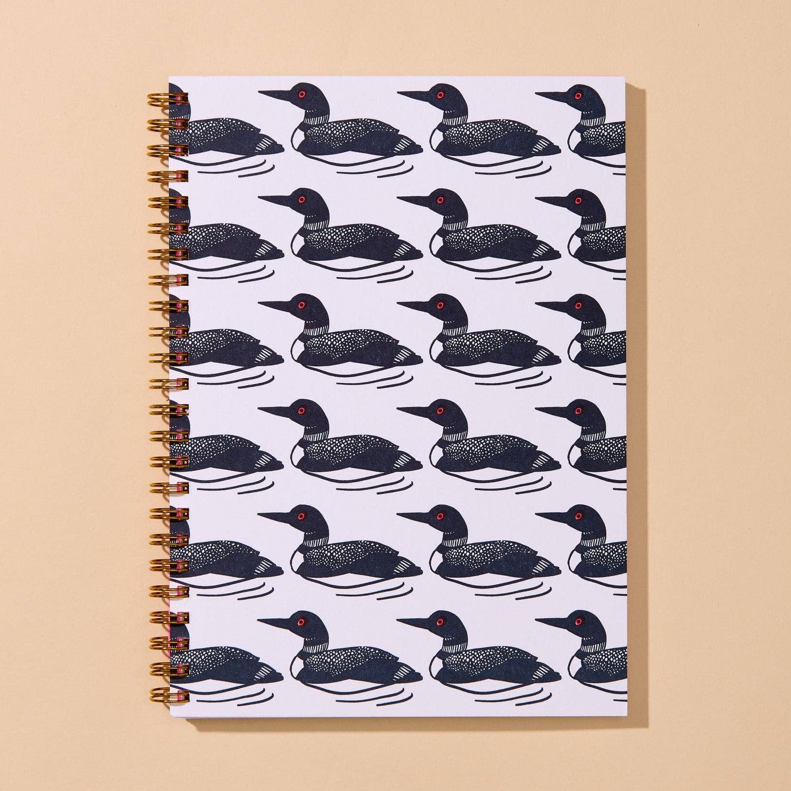 A spiral-bound notebook with a white cover printed with a repeating black-and-white duck pattern, featuring a bright pink front cover with red text reading 'NOTE BOOK' and smaller text below. The notebook is photographed on a warm beige background.