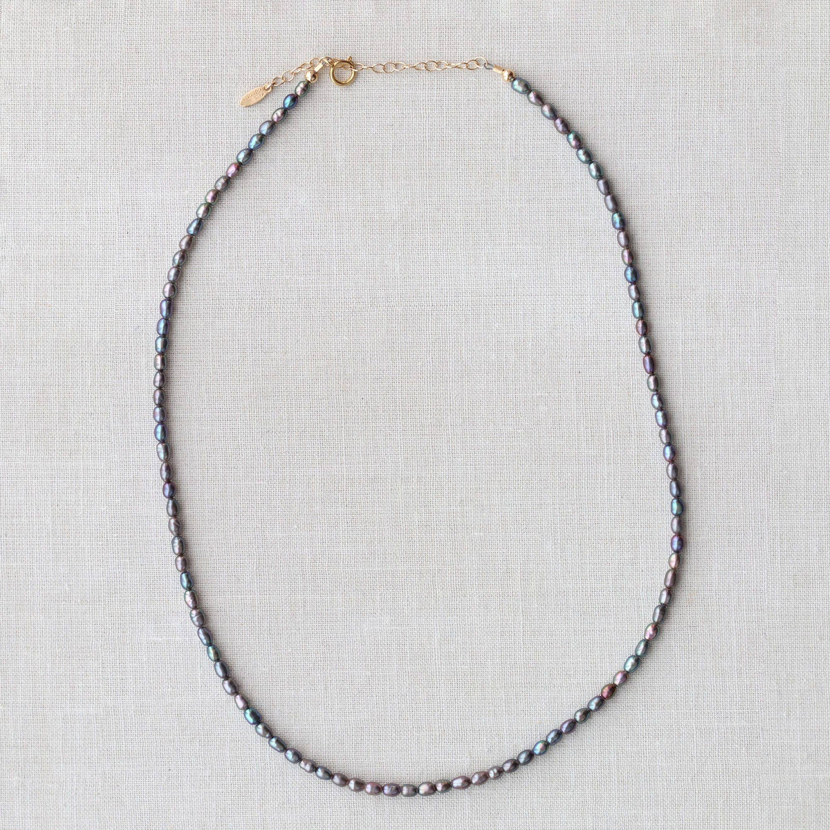 A beaded necklace made of small iridescent gray-blue pearls with gold-toned chain links and extender, laid flat on a light beige linen background.