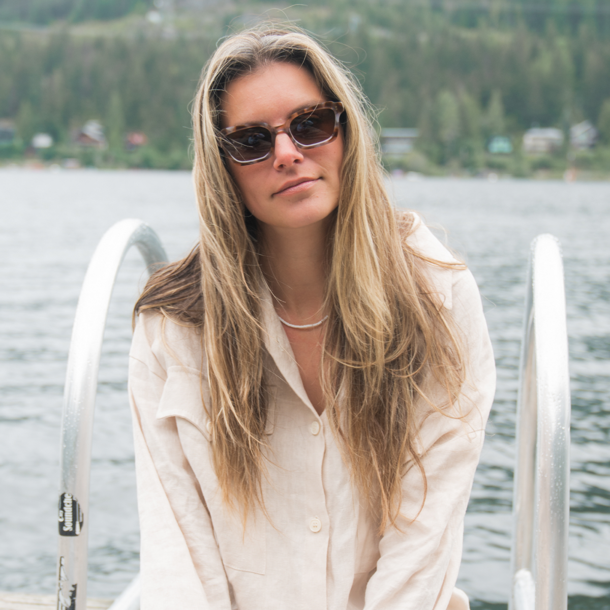 Woman sitting on a dock by a lake wearing sunglasses and a light-colored outfit.
