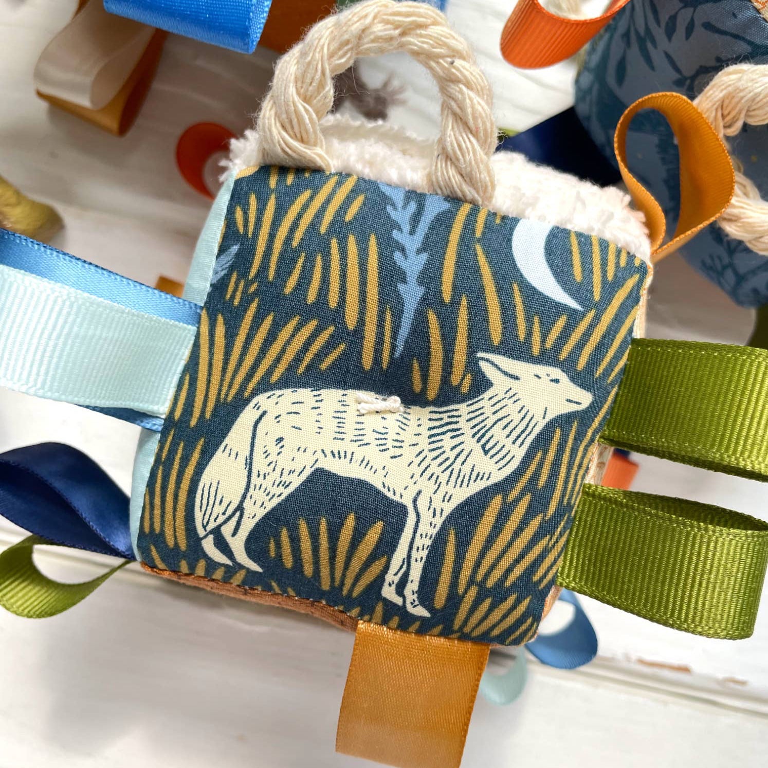 A baby sensory taggie block featuring a wolf design in blue, gold, and green fabrics with ribbons and a rattle.