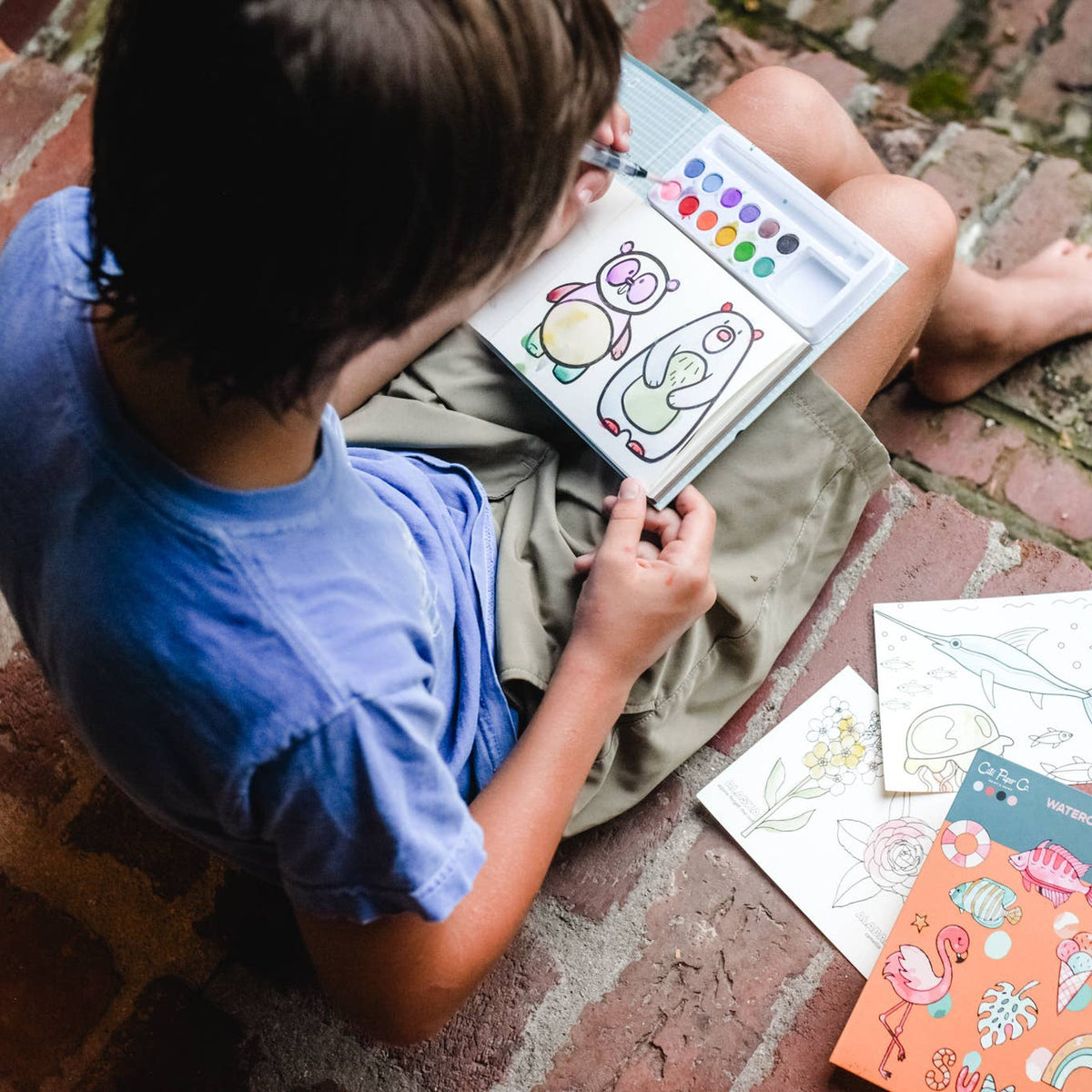 Child coloring a picture on a brick floor with coloring books around