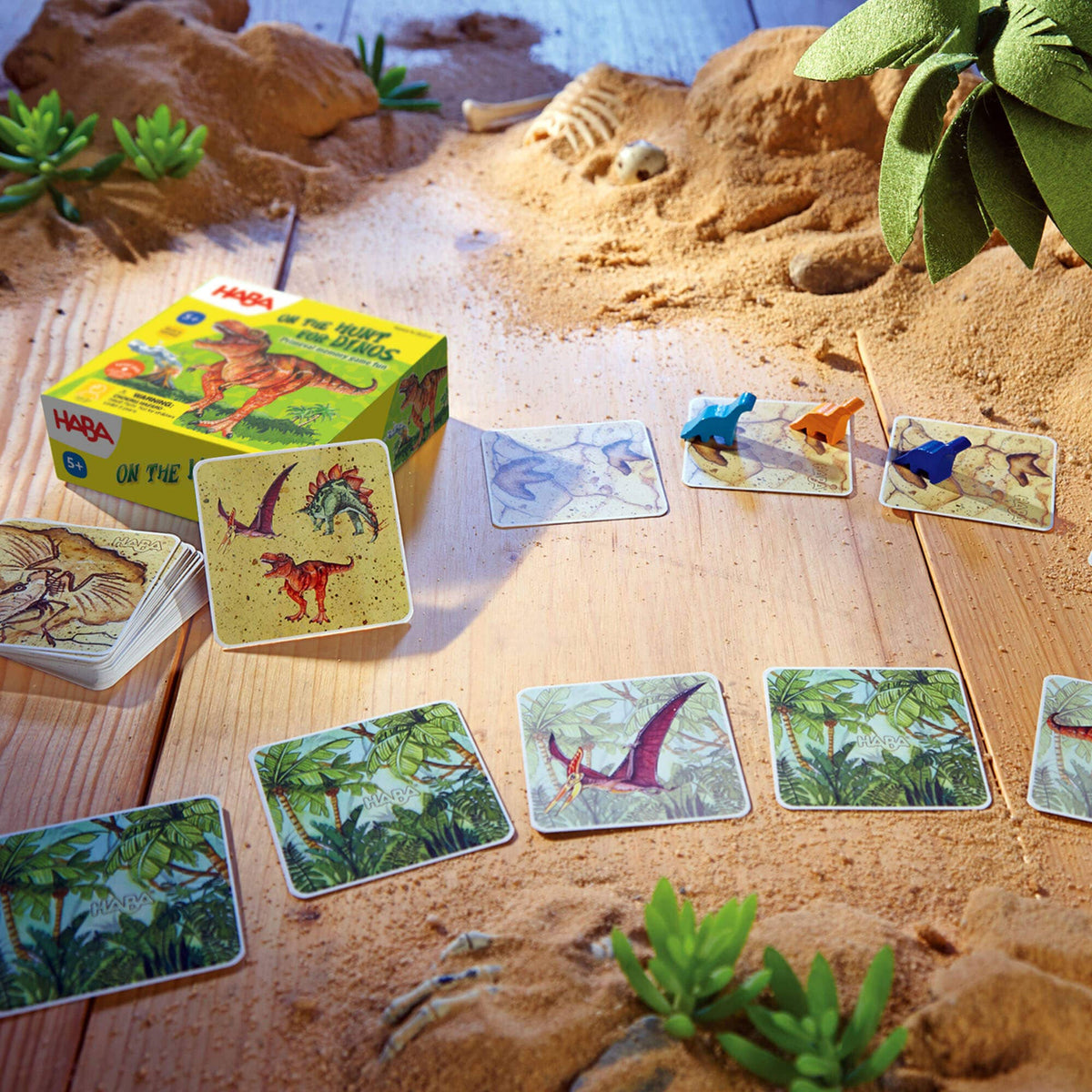 Dinosaur-themed card game on a wooden surface with sand and plants