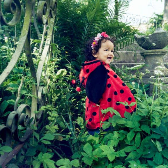 Child in a garden wearing a red and black outfit with polka dots, surrounded by greenery.