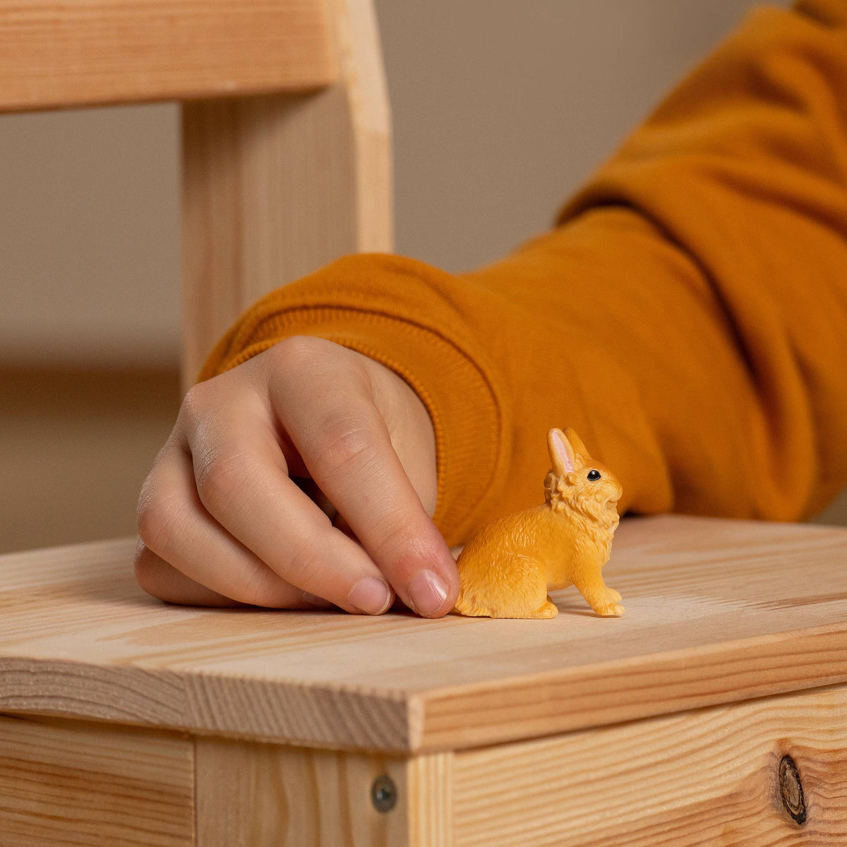 A small yellow rabbit figurine sits on a light wood surface, with a person's hand resting near it and a mustard-orange sleeve visible in the background.