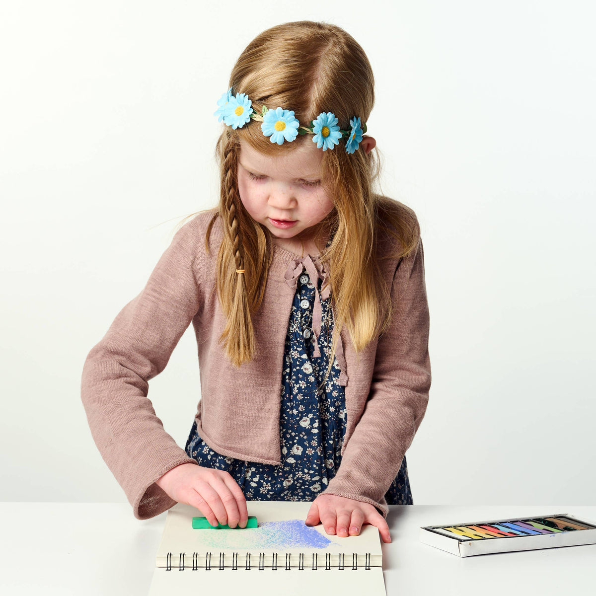 Young girl with a flower headband playing with pastels on a white background