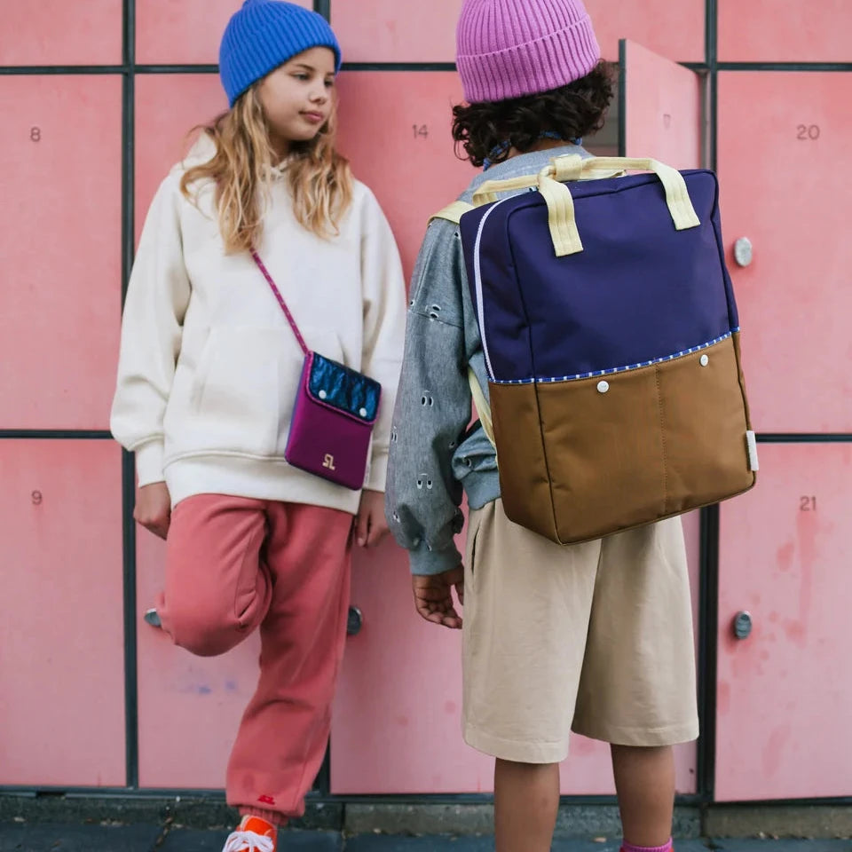 Two children standing in front of pink school lockers, one wearing a blue beanie, cream sweatshirt, and coral pants with a purple crossbody bag, and the other wearing a pink beanie, gray shirt, beige shorts, and a navy and brown backpack.