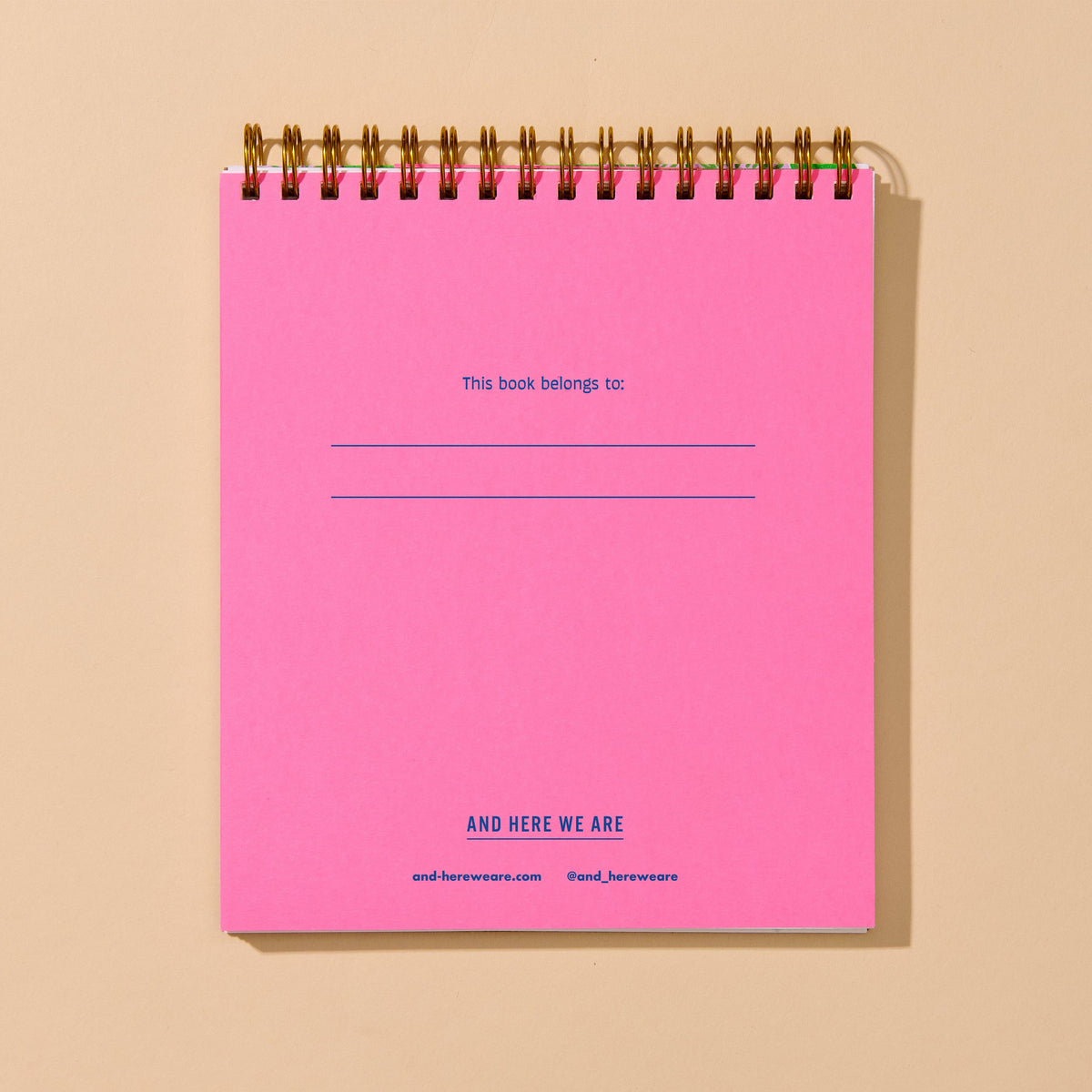 A bright pink spiral-bound notebook with blue printed text, shown standing upright against a warm beige background.